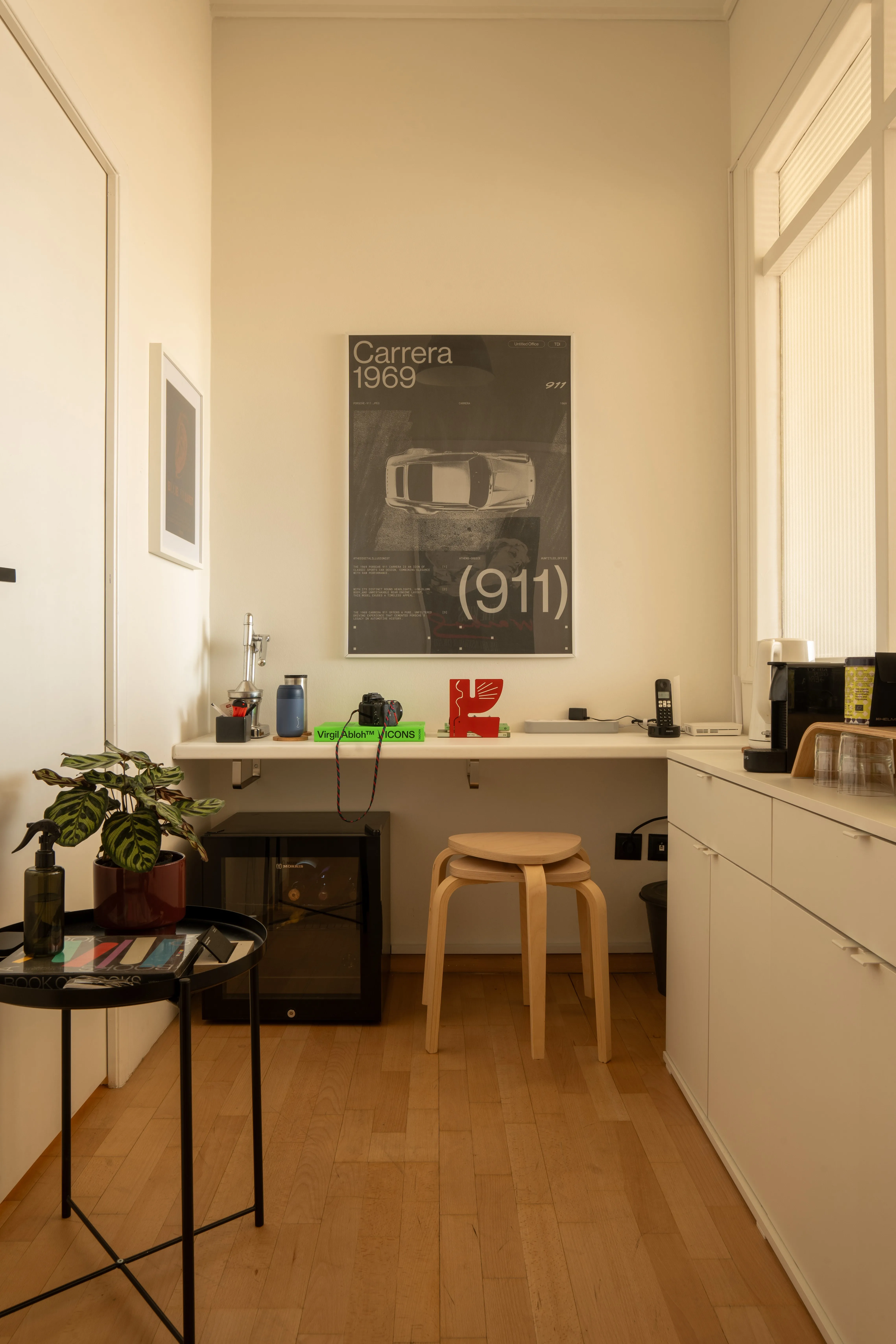 A corner of the studio: a Carrera 1969 poster above a narrow desk, a wooden stool and a plant on a side table