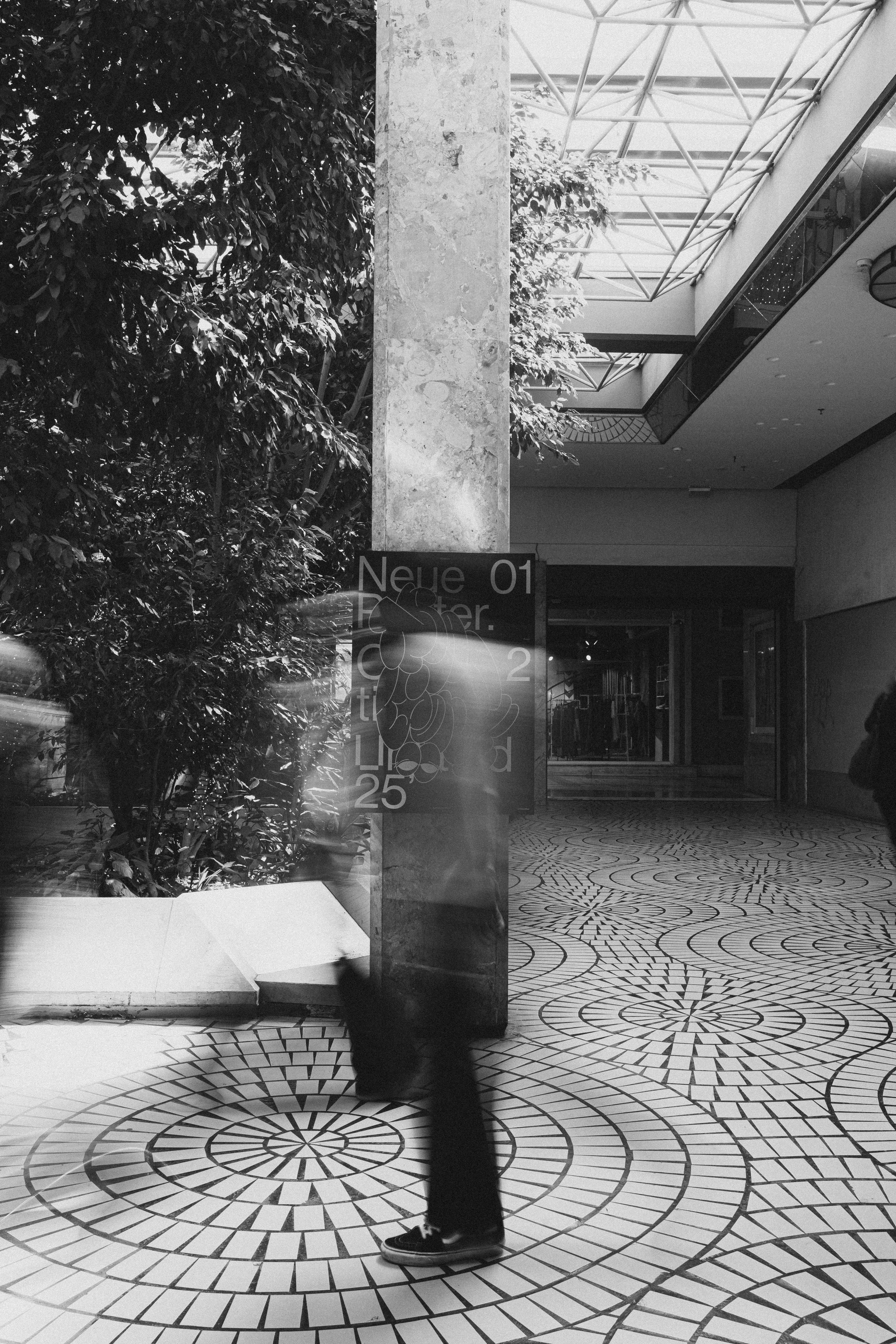 Black-and-white arcade with a marble column, a circular mosaic floor and a motion-blurred passer-by in front of a poster