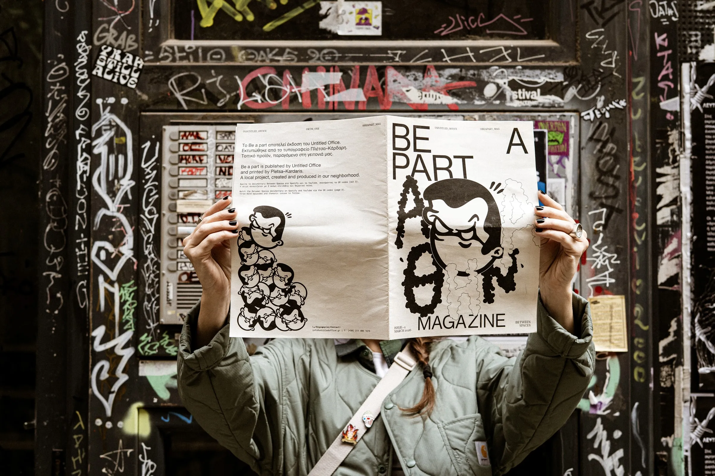 A person holding up the Be A Part magazine in front of their face against a graffiti-covered wall