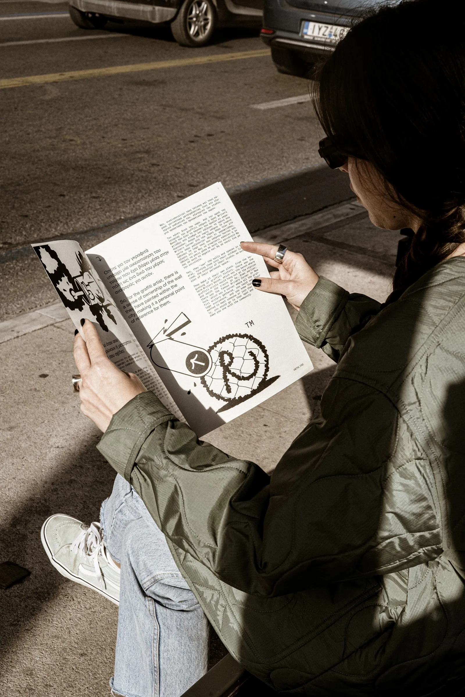 A reader sitting on the pavement with the magazine open, parked cars behind