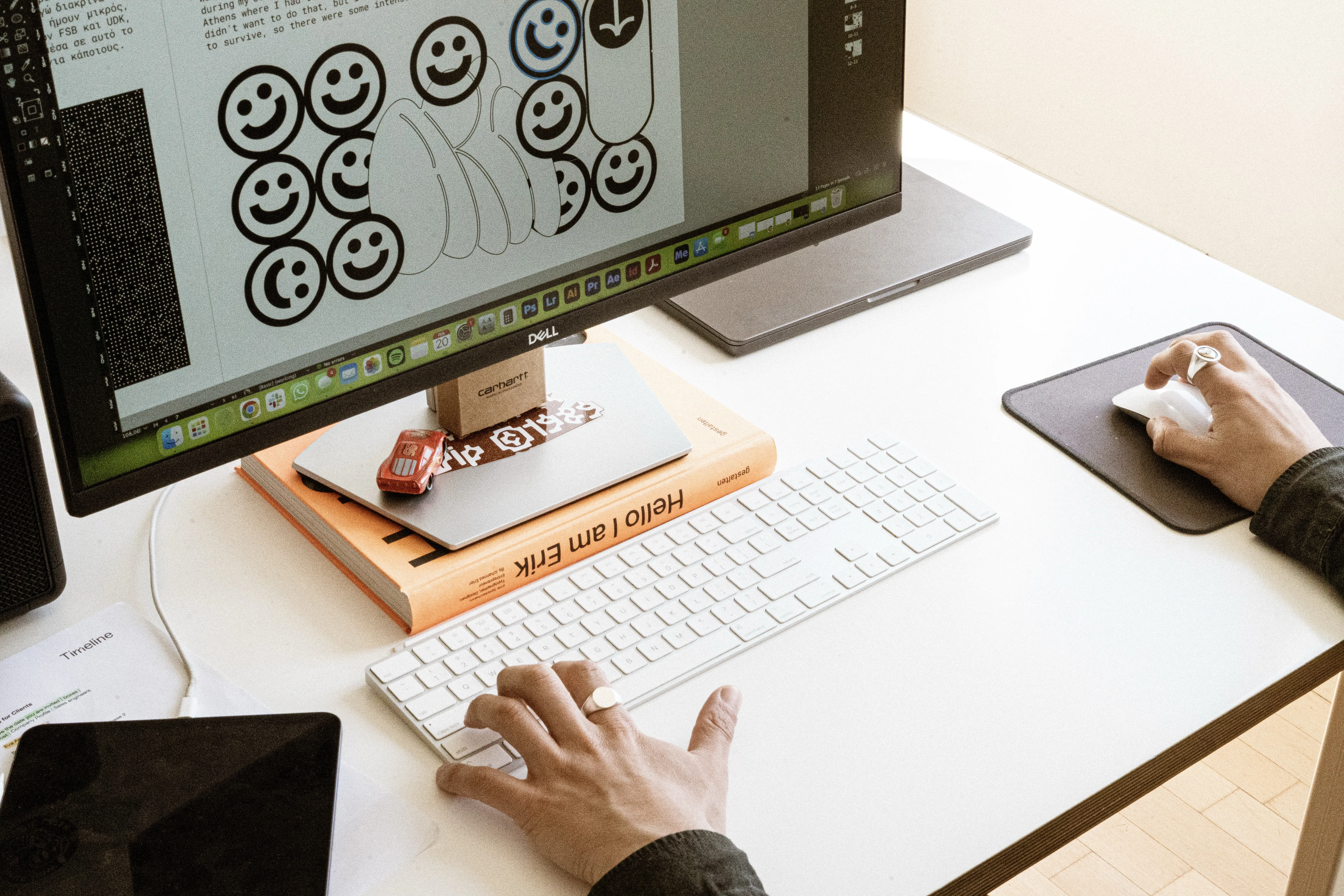 Hands at a white keyboard and mouse on a studio desk, below a monitor showing a page of smiley-face illustrations and Greek text