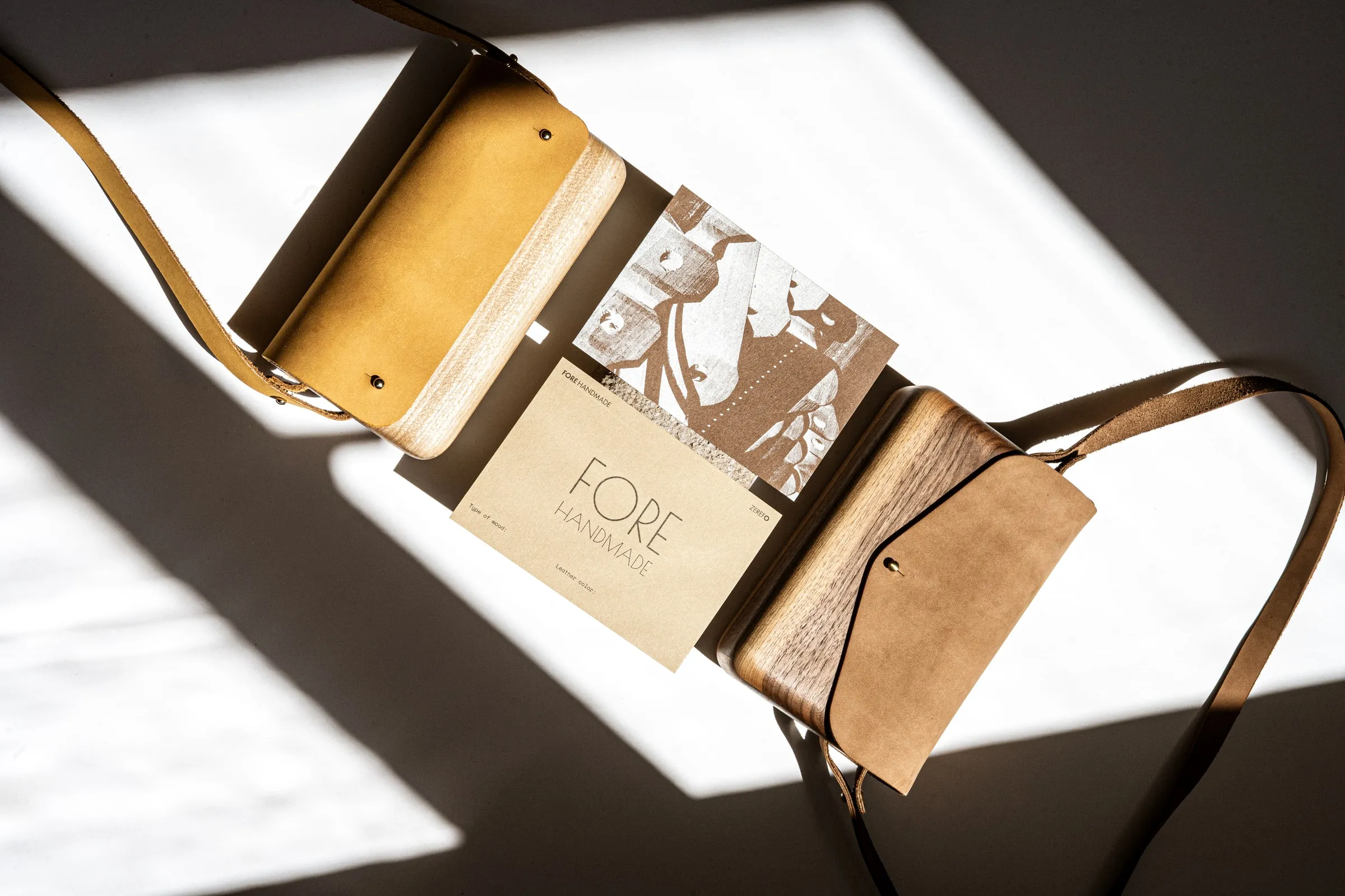 Two wooden bags with leather straps on either side of a FORE HANDMADE card, in hard sunlight
