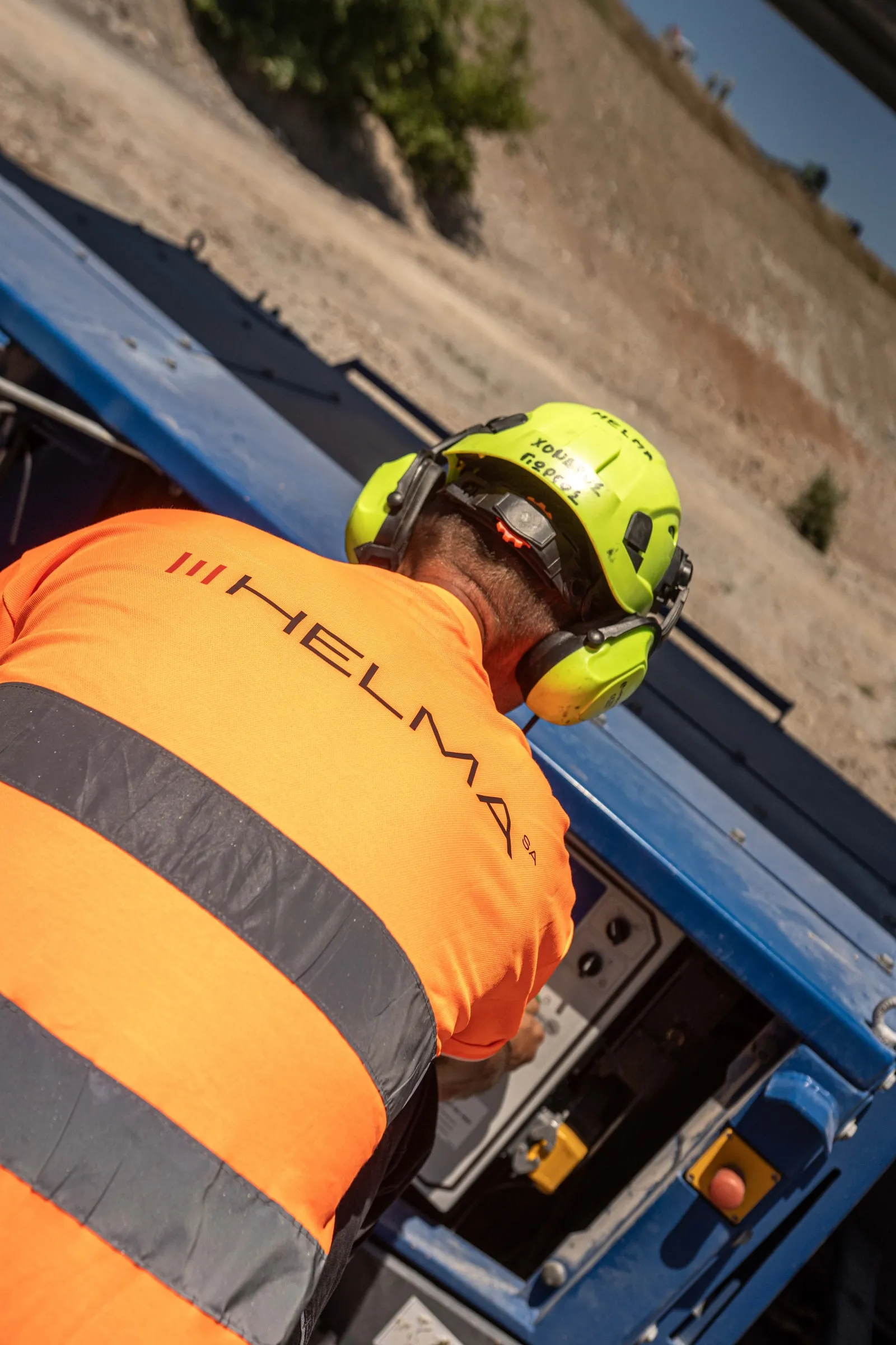 A worker in an orange hi-vis jacket with the Helma logo and yellow ear defenders at machine controls