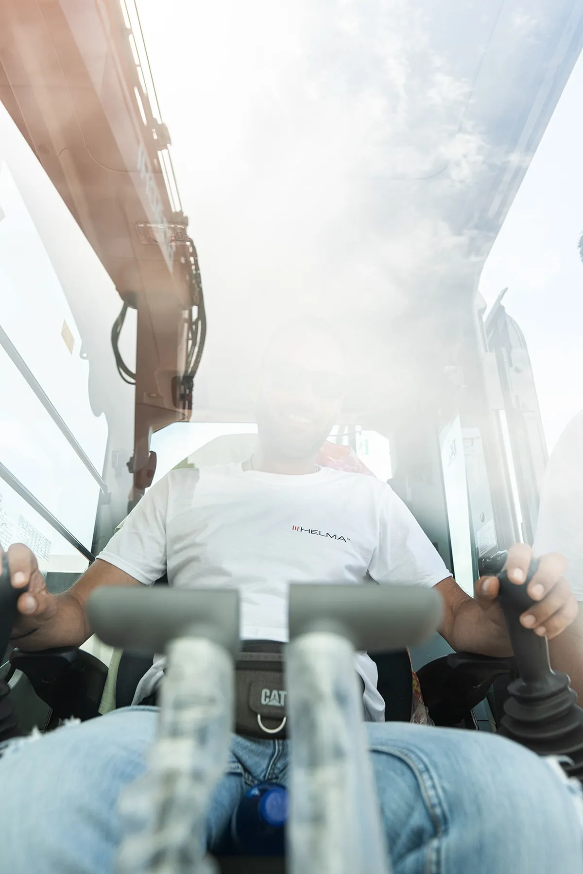 An operator in a white Helma t-shirt at the controls inside an excavator cab