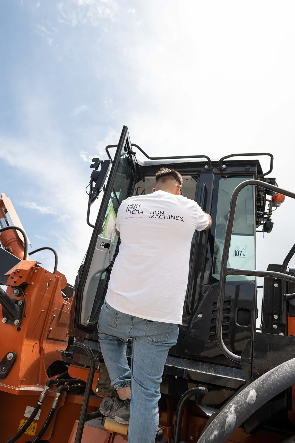 A person in a white NEXT GEN→ERA·TION MACHINES t-shirt climbing into an orange machine, seen from behind