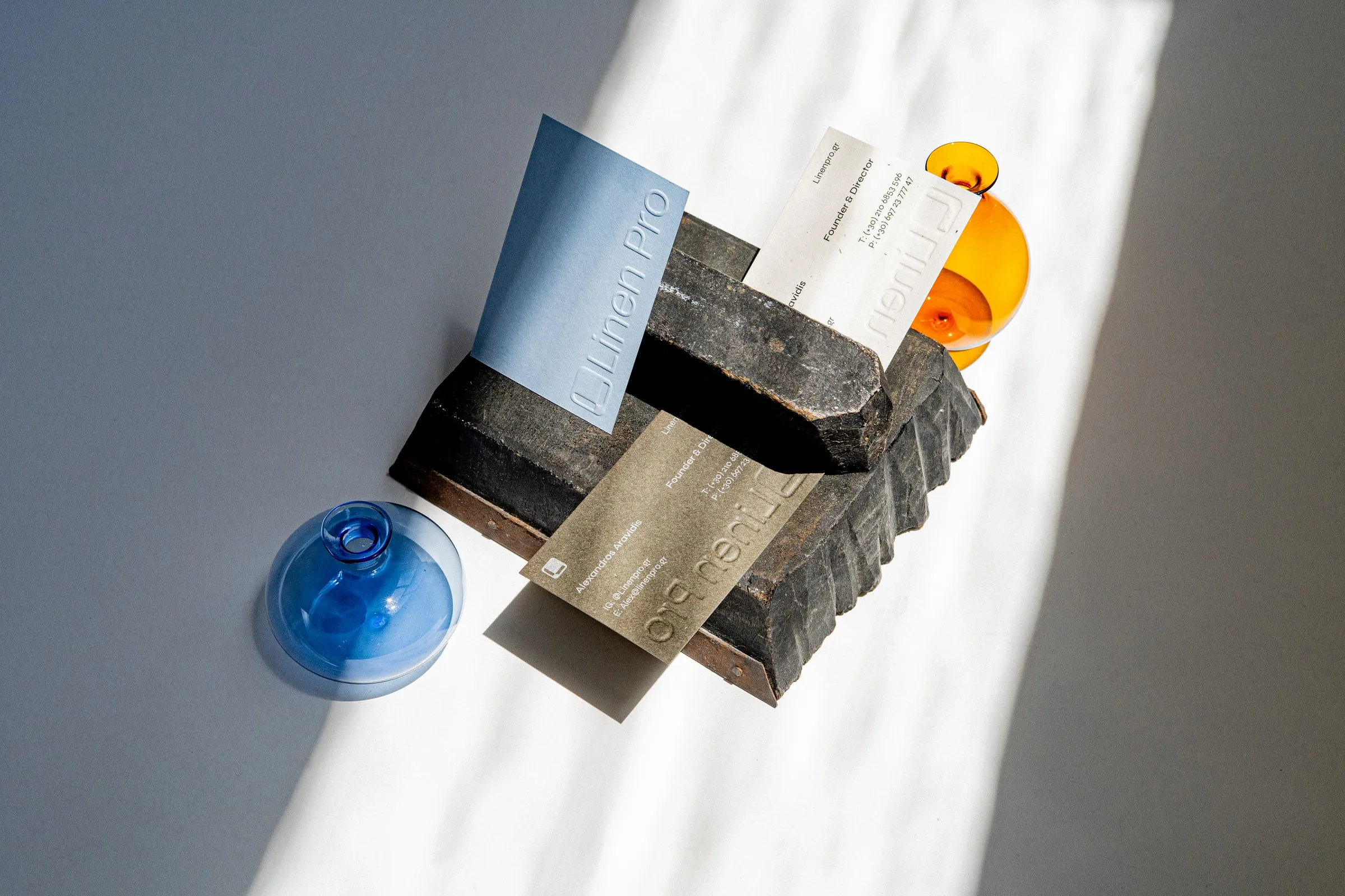 Pale blue and kraft Linen Pro cards on a dark brick between blue and amber glass globes, in a band of sunlight