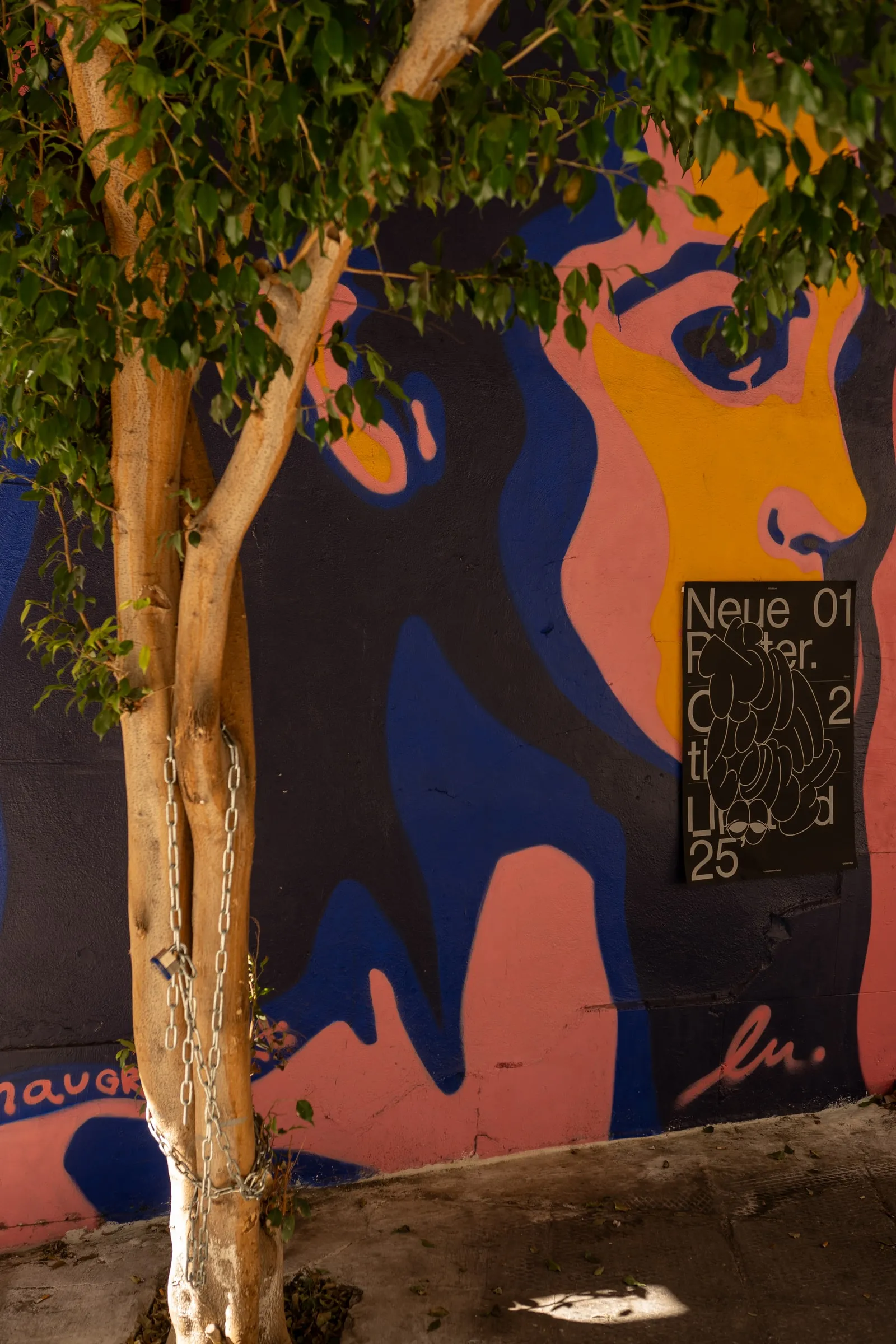 A small copy of the poster on a large mural of a woman's face, behind a tree