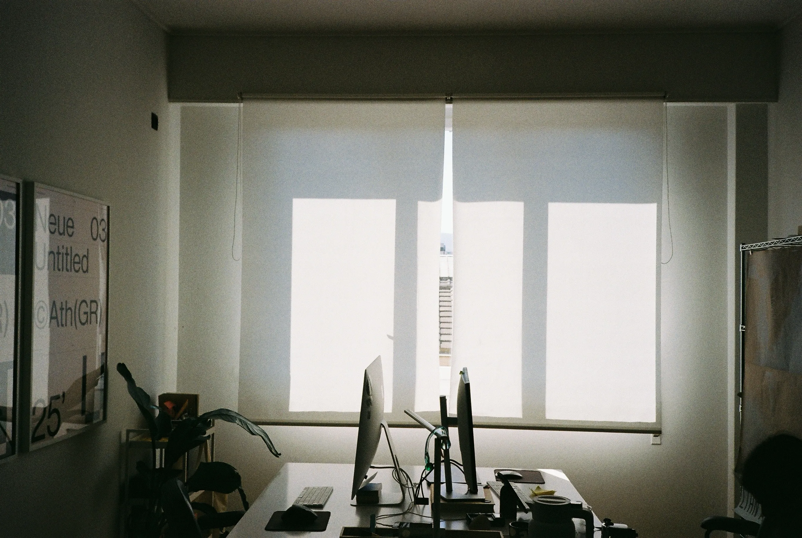 Two monitors back to back on a shared desk in front of a sunlit roller blind