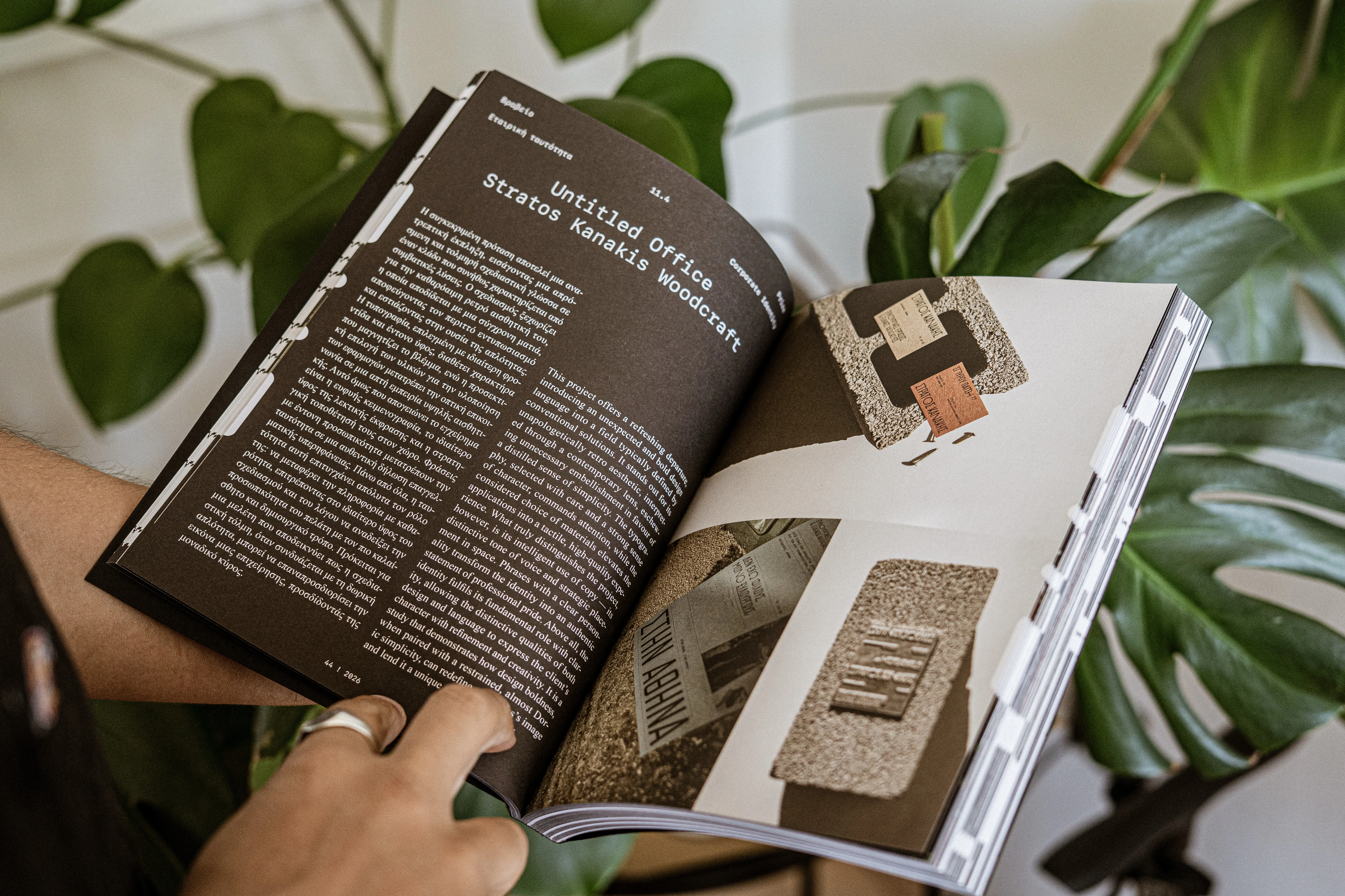 Hands holding open a magazine spread about a corporate identity project, with monstera leaves behind