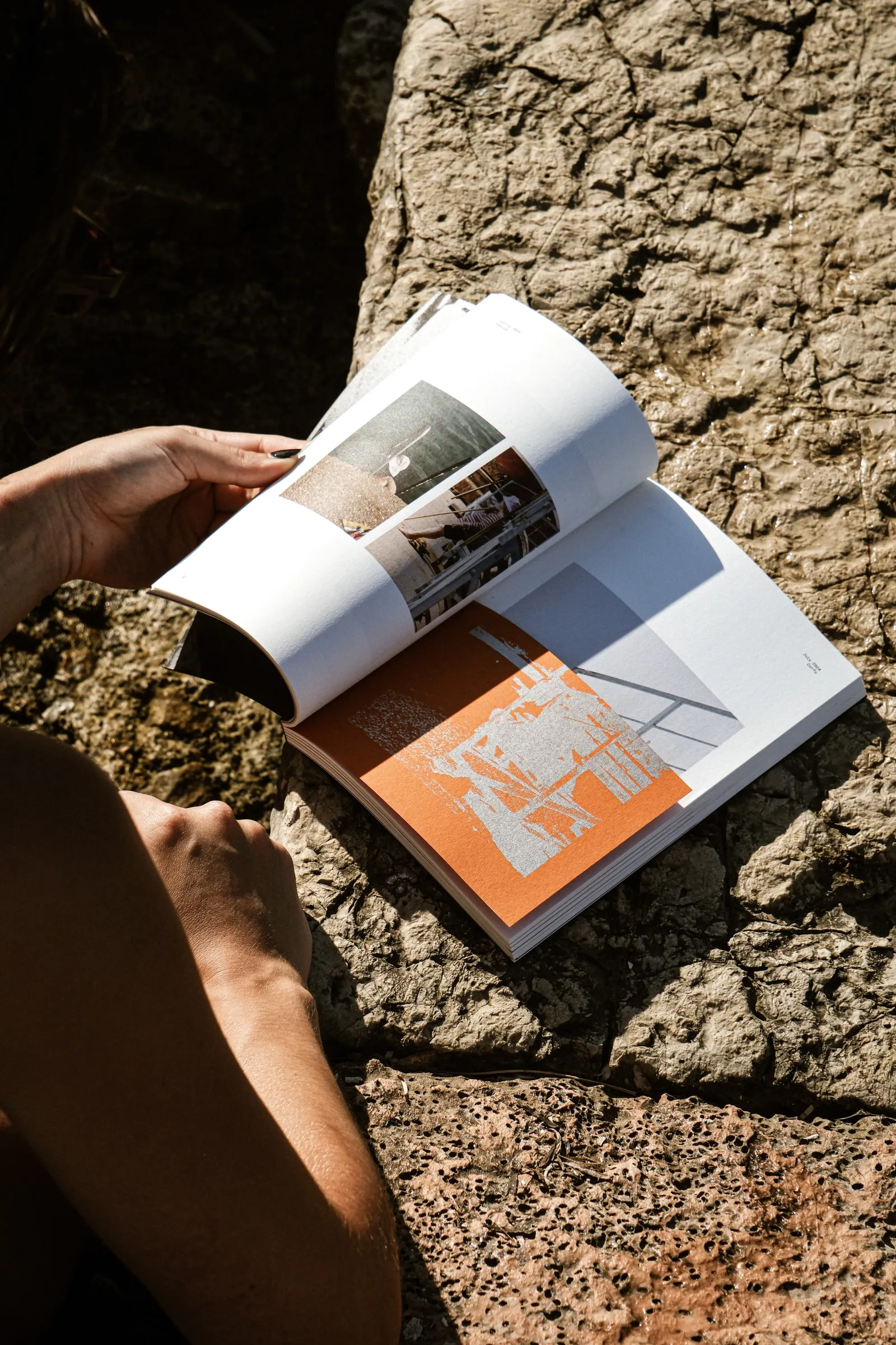The photobook open at an orange-tinted spread on warm stone, a hand holding the page