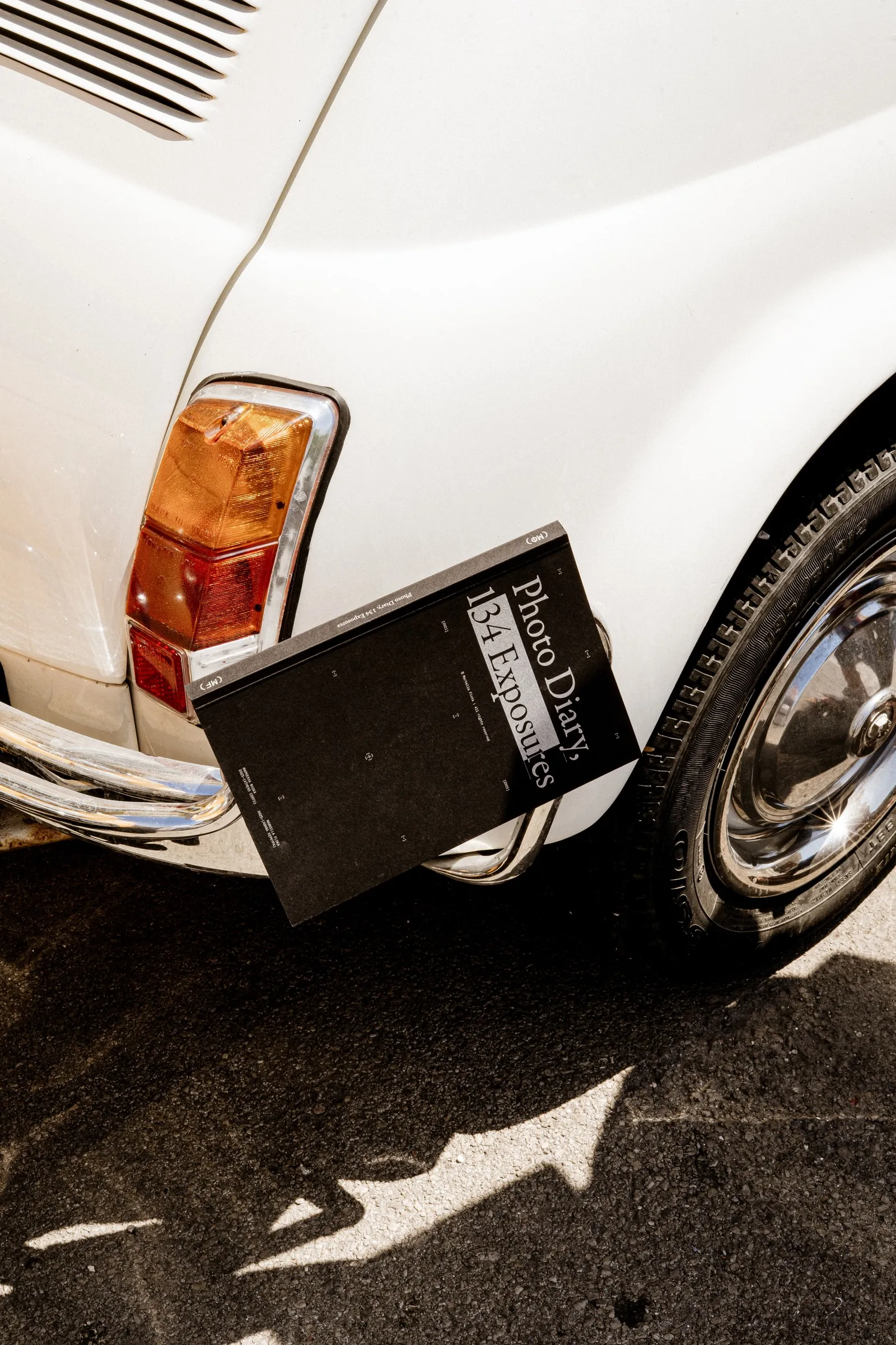 The black photobook leaning against the rear of a white vintage car