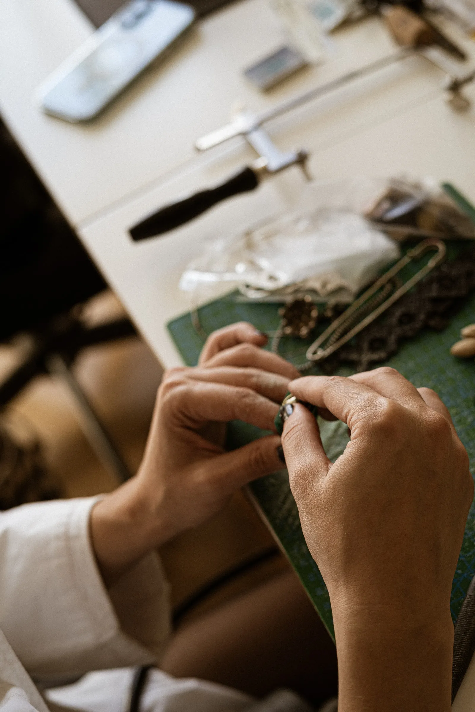 Close-up of hands working on a small metal piece over a green cutting mat, jewellery tools on the desk behind