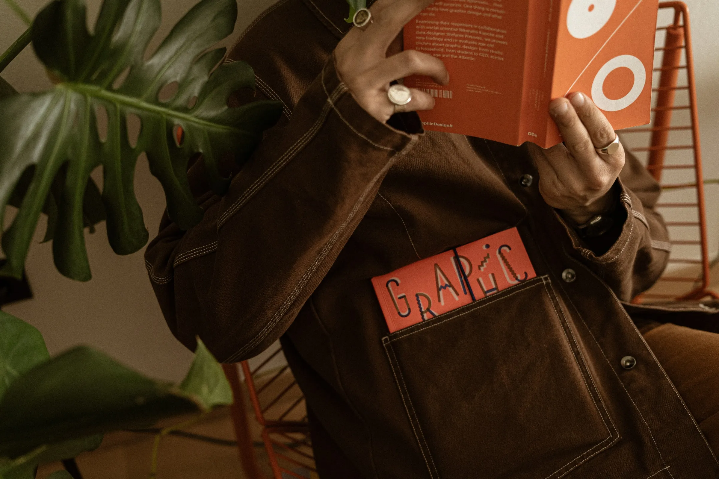 A person in a brown work jacket reading an orange book held up in front of their face, another book in their pocket, beside a monstera