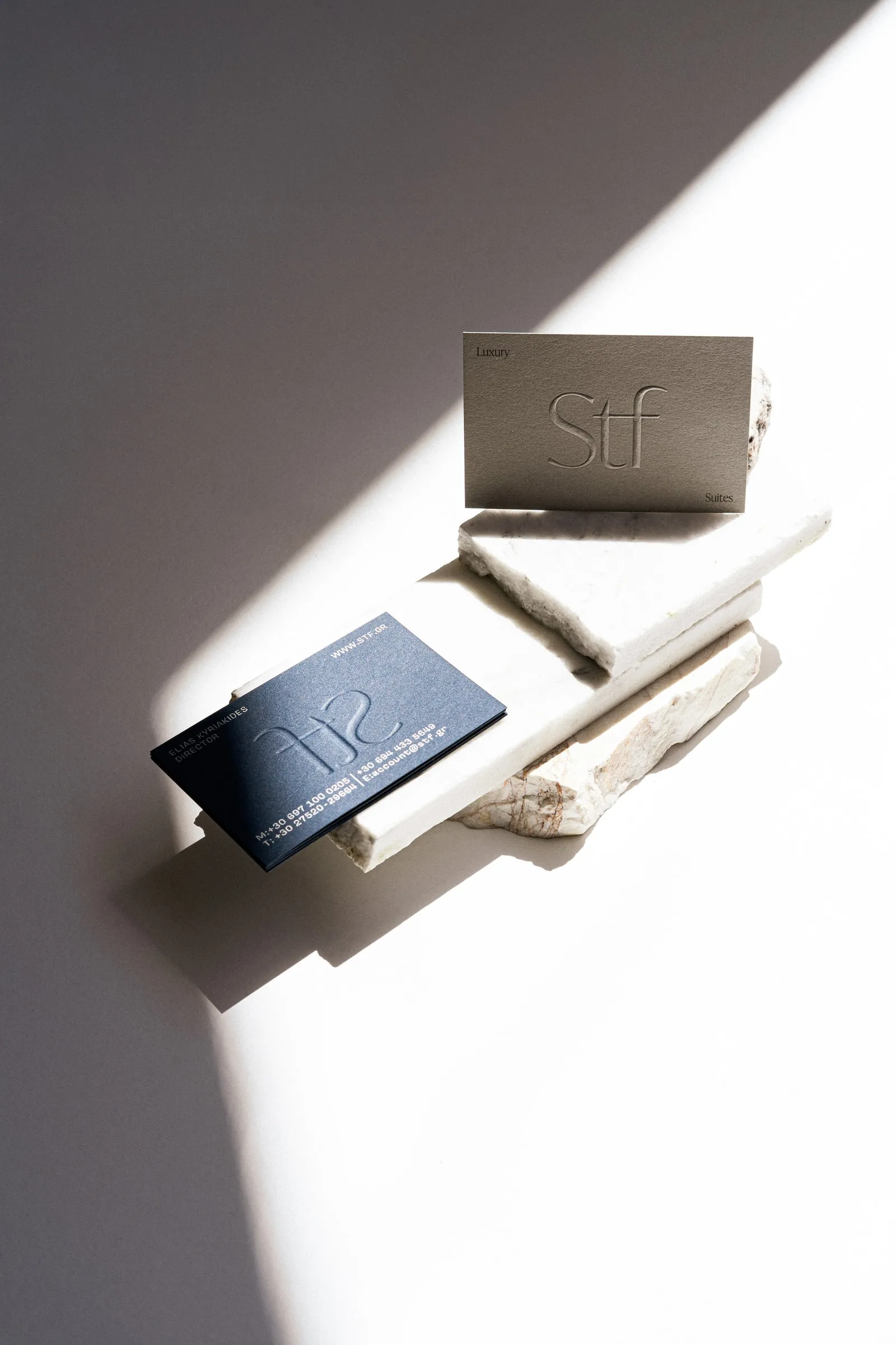 Embossed STF cards in clay and navy on stacked white marble slabs, in raking sunlight