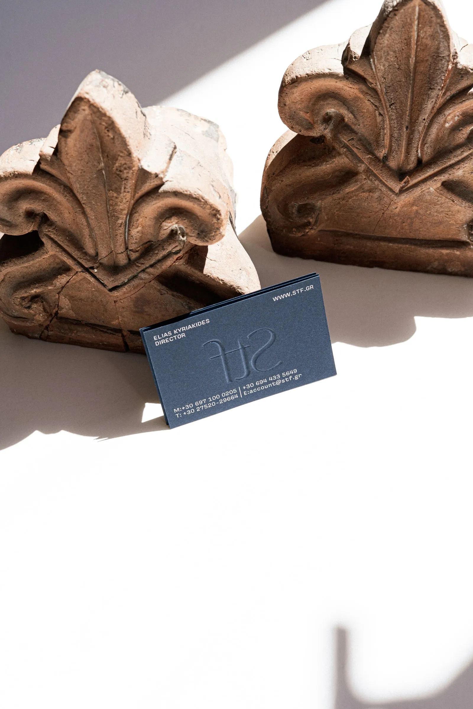 A navy business card with a mirrored embossed STF monogram, leaning against two terracotta ornaments
