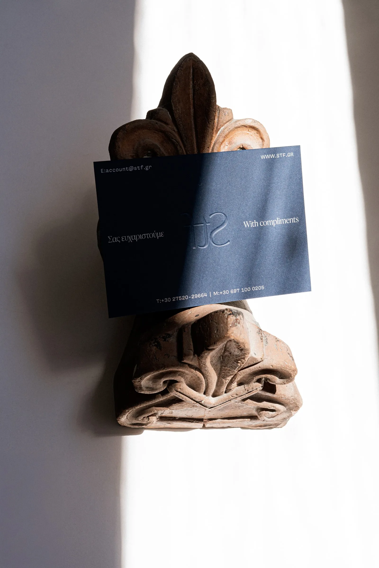 A navy With compliments card with an embossed STF monogram, tucked into a terracotta ornament on a sunlit wall