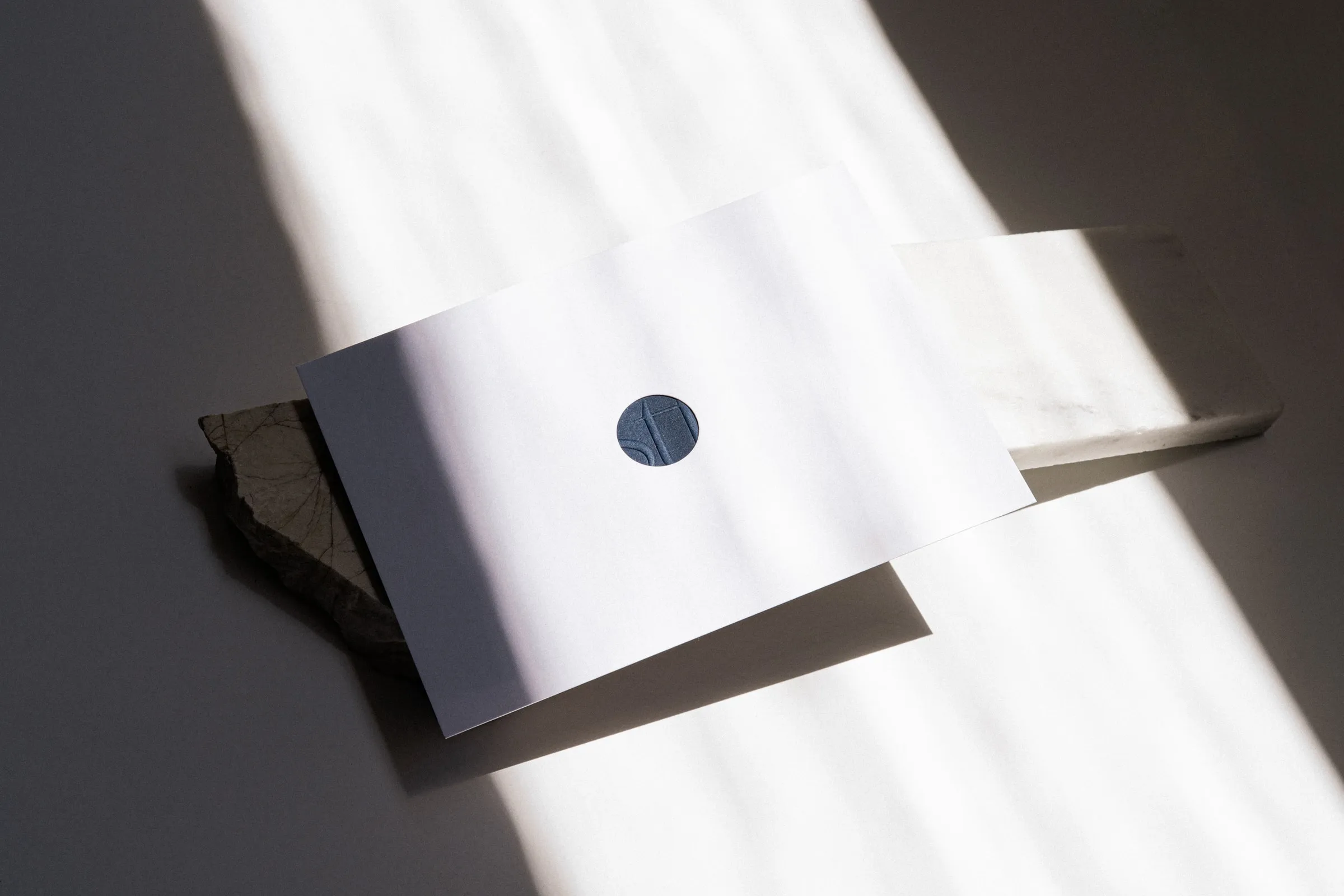 A white envelope with a die-cut circle revealing the embossed navy STF logo underneath, resting on marble
