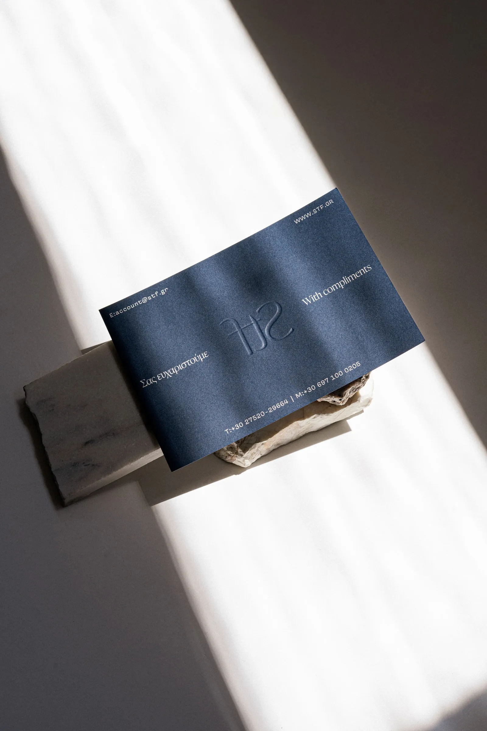 A navy With compliments card with an embossed STF monogram, resting on marble in a band of sunlight