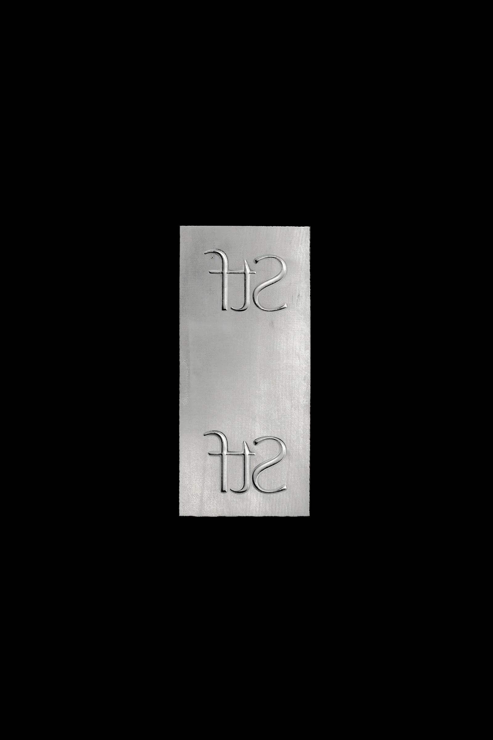 The aluminium embossing plate with two mirrored STF monograms, photographed on black