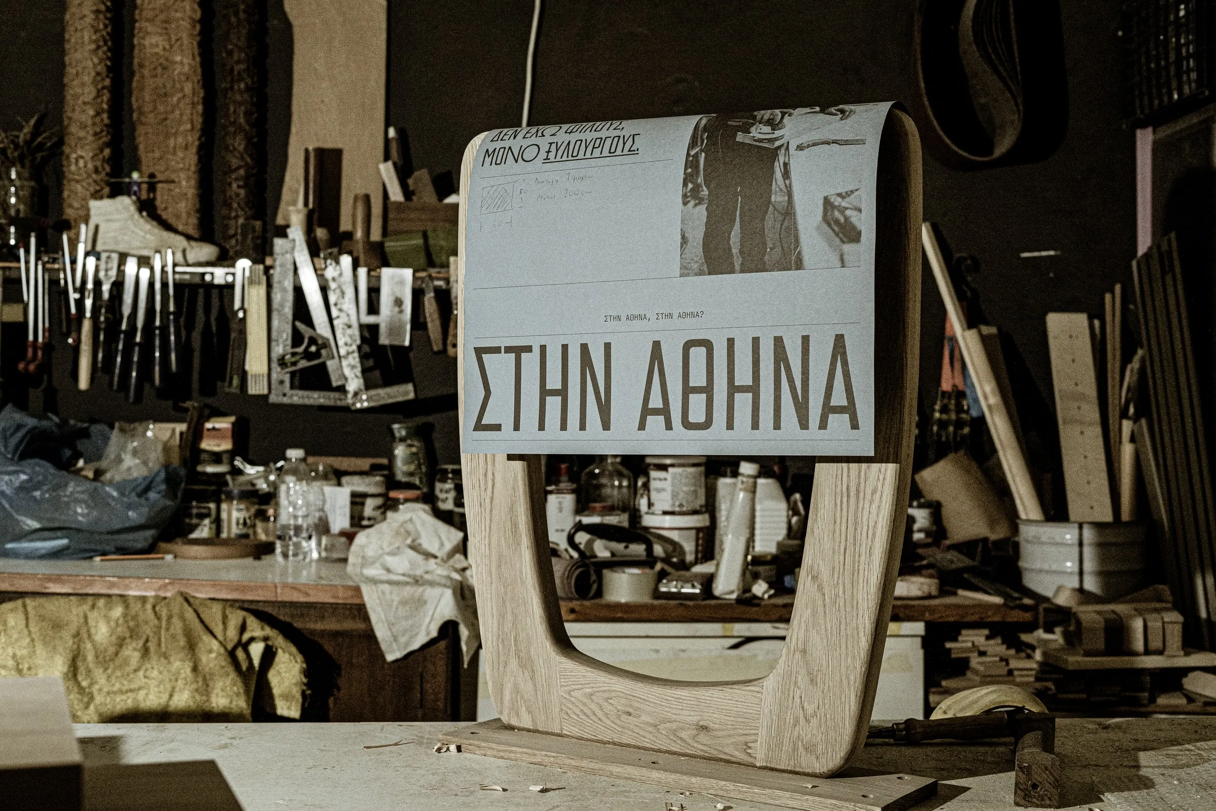 A grey poster reading ΣΤΗΝ ΑΘΗΝΑ draped over a curved wooden frame in a cluttered carpentry workshop