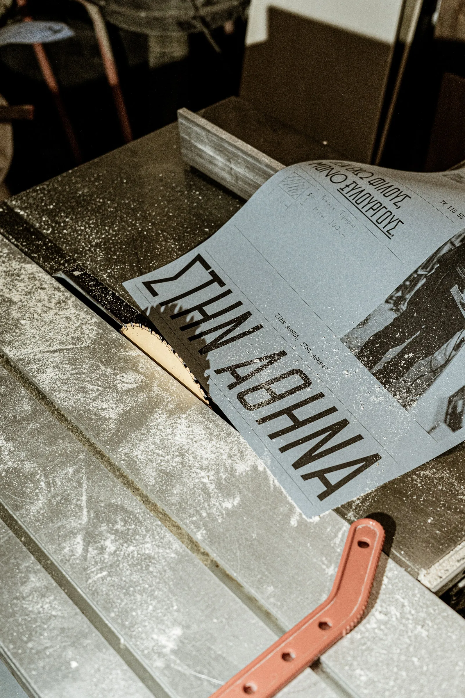 A ΣΤΗΝ ΑΘΗΝΑ poster lying on a circular saw table dusted with sawdust