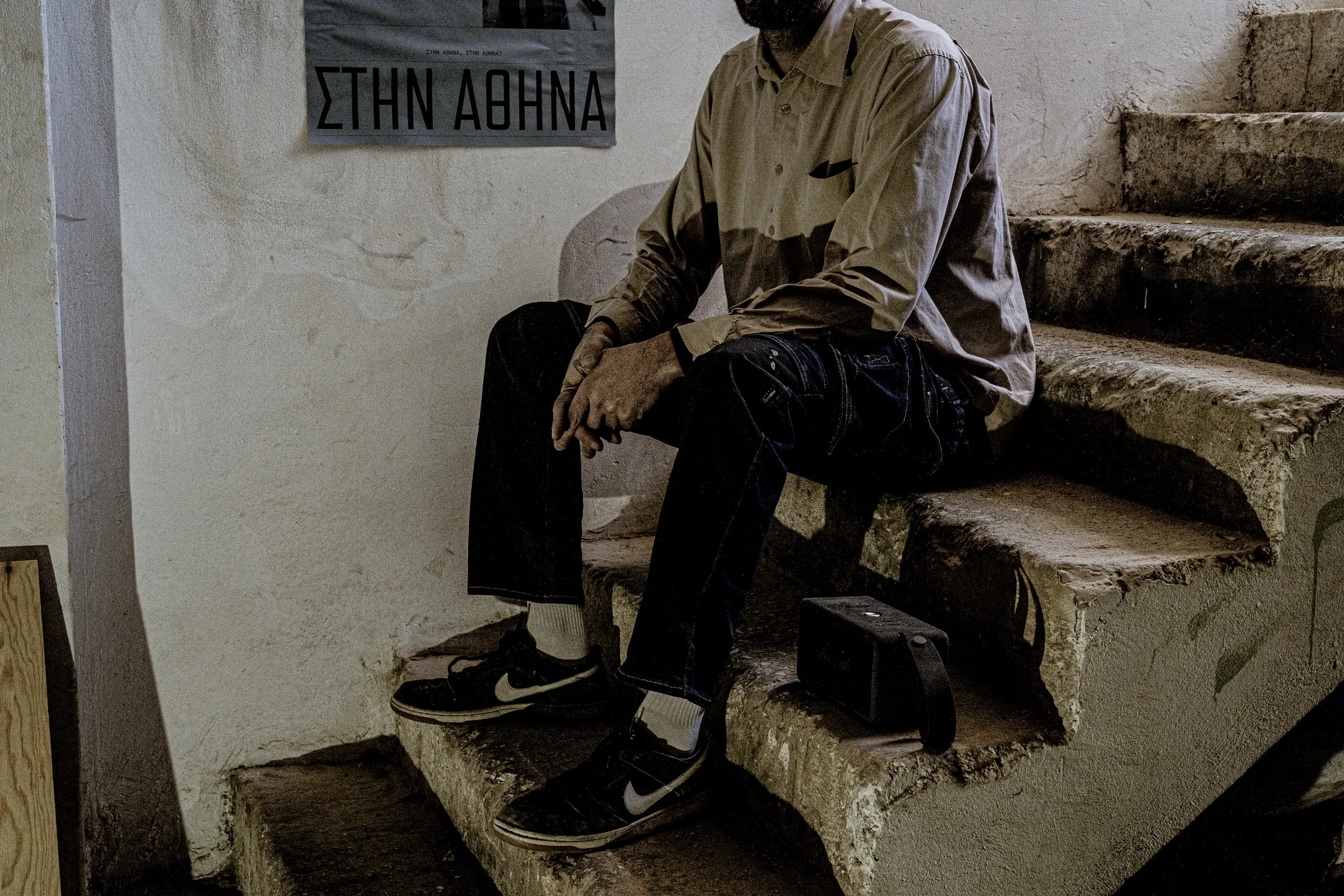 A person sitting on worn concrete steps below a ΣΤΗΝ ΑΘΗΝΑ poster on the wall
