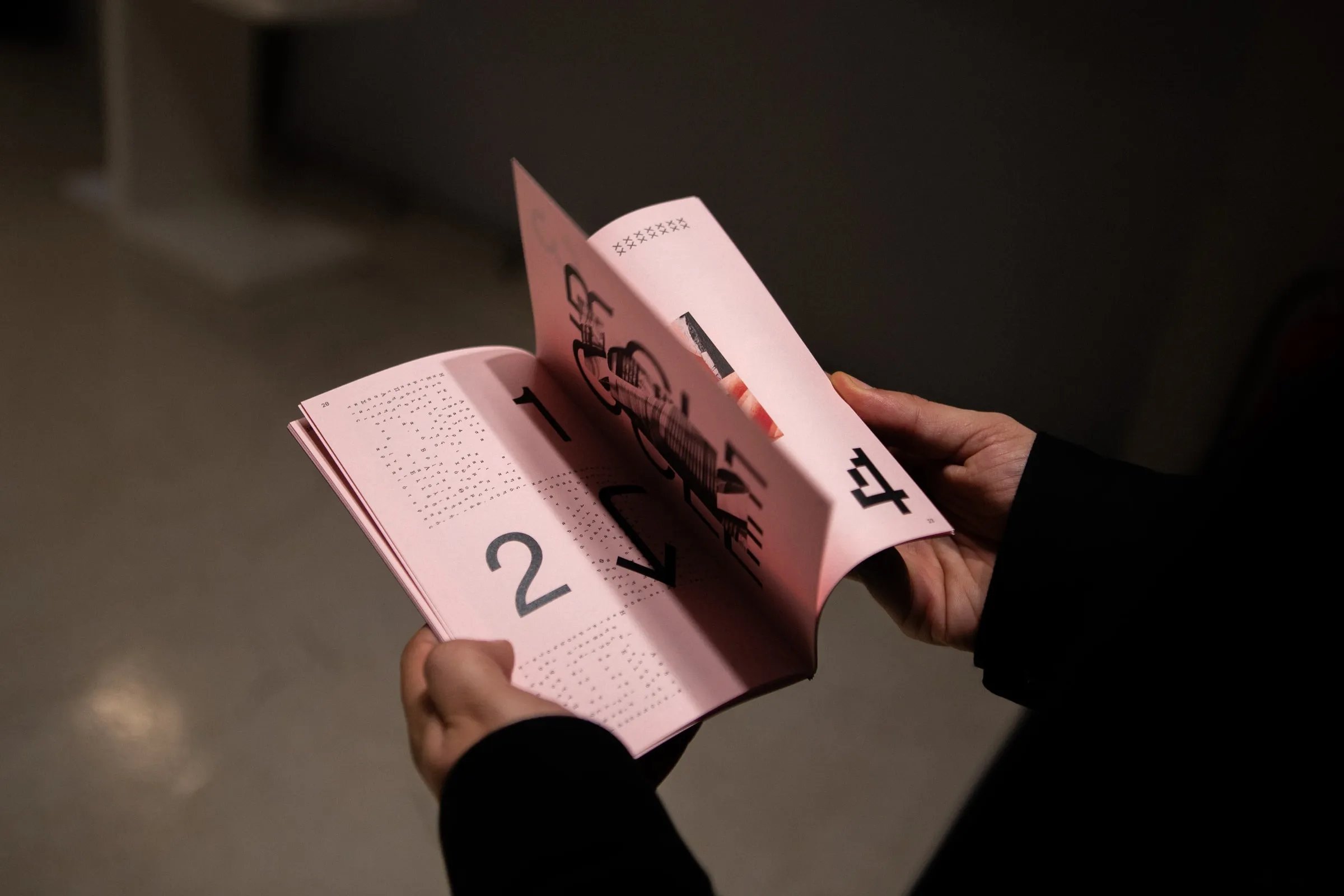 Hands holding the zine open at a pink spread in a dim gallery