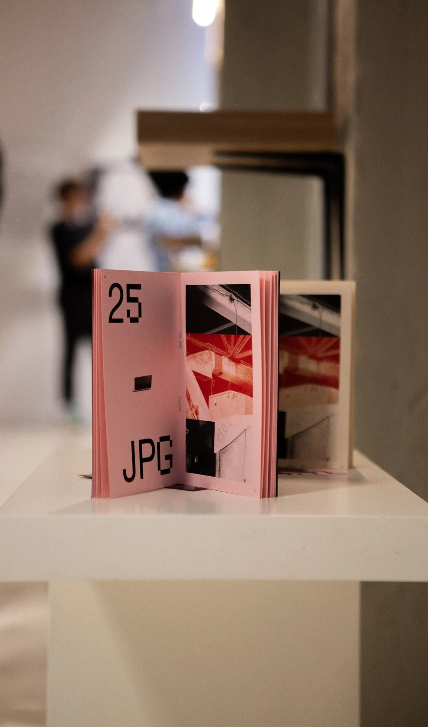 The zine standing open on a white plinth at the exhibition, visitors blurred behind