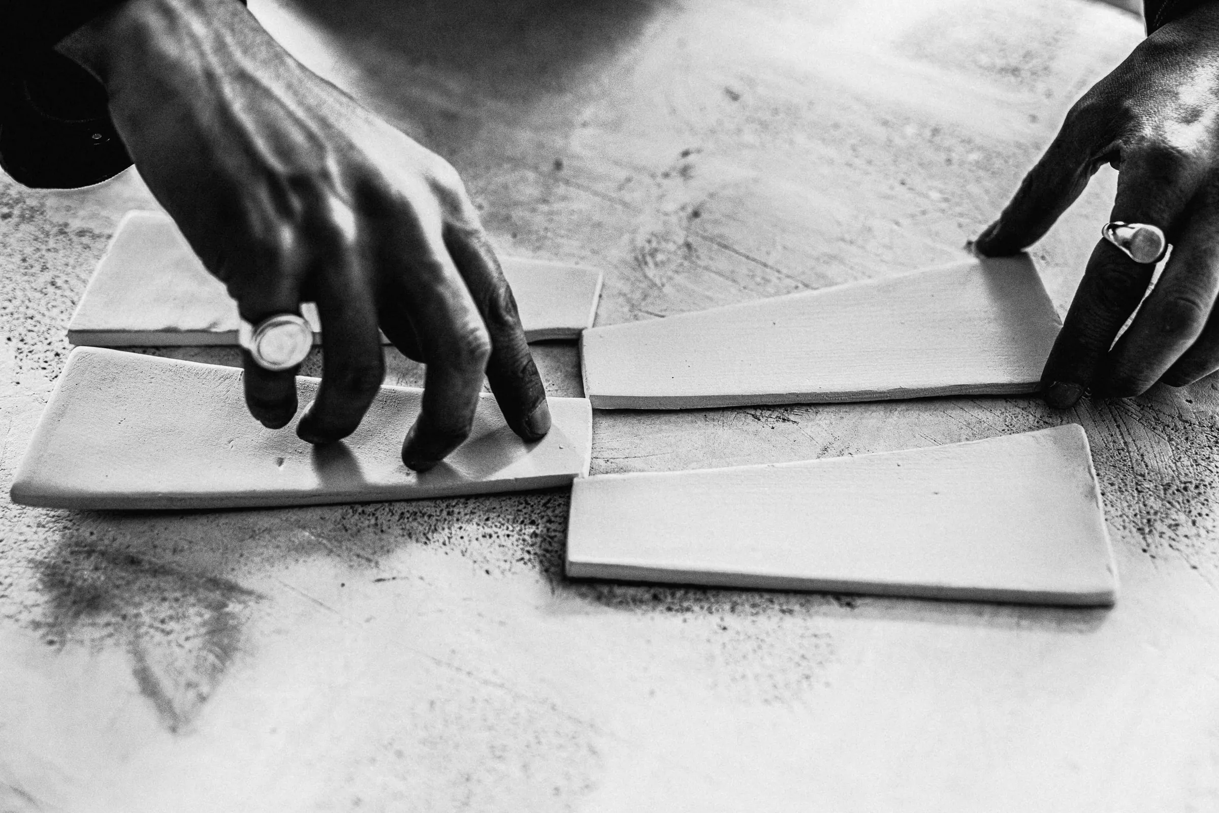 Black-and-white close-up of hands arranging flat clay slabs on a worktable