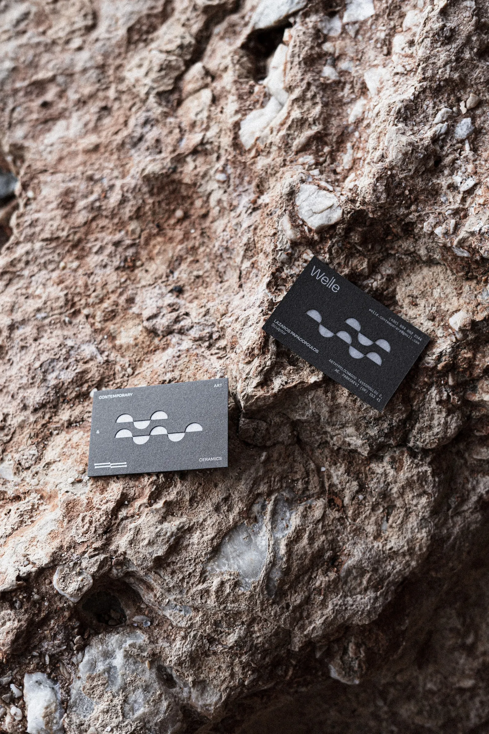 Two dark grey Welle business cards with a die-cut wave pattern, lying on red rock