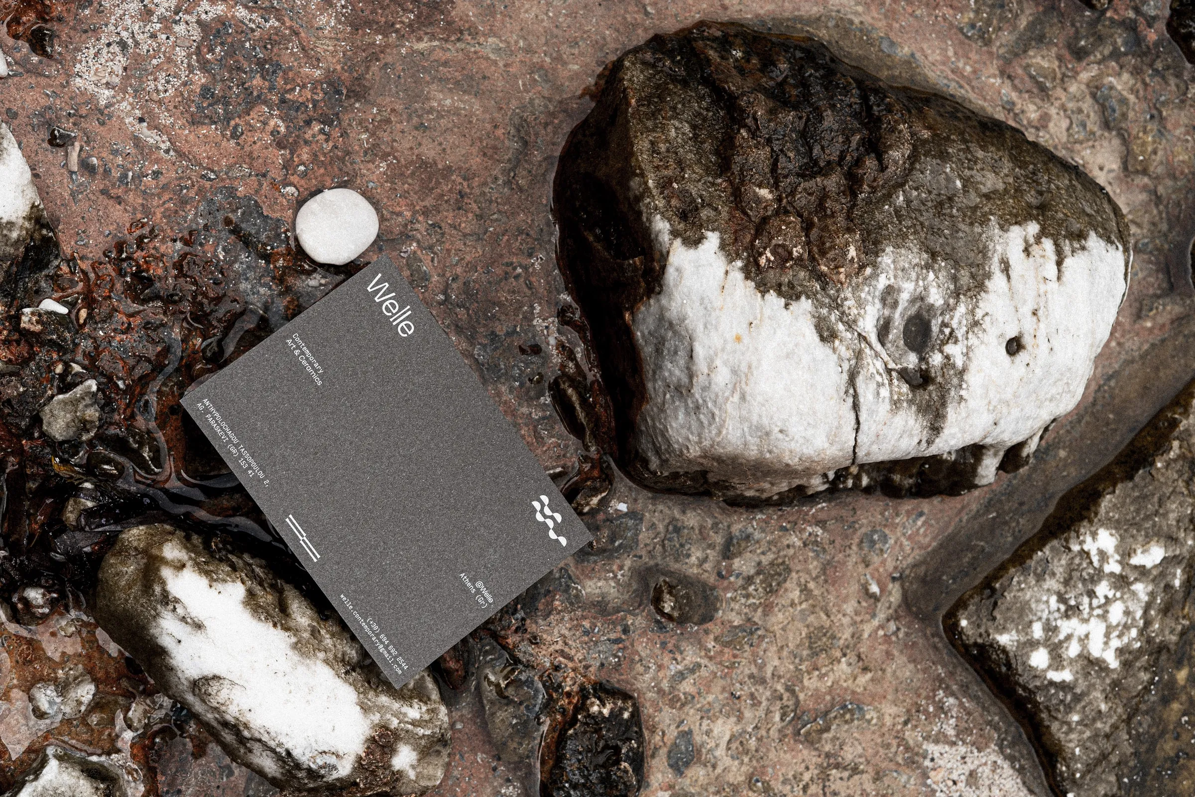 A dark grey Welle card resting on wet rocks beside a large white-veined stone