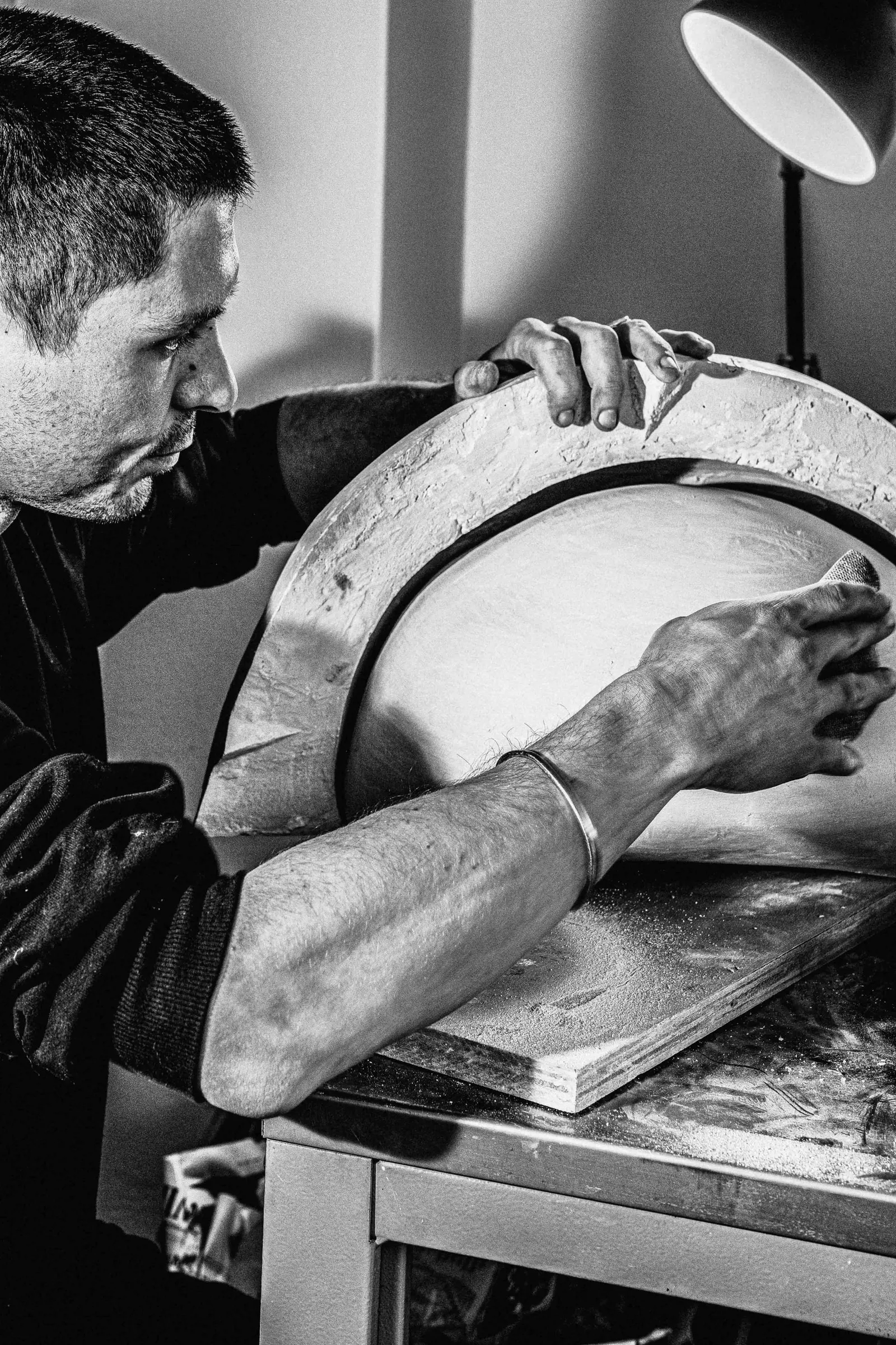 Black-and-white photo of a sculptor sanding a large curved plaster mould under a desk lamp