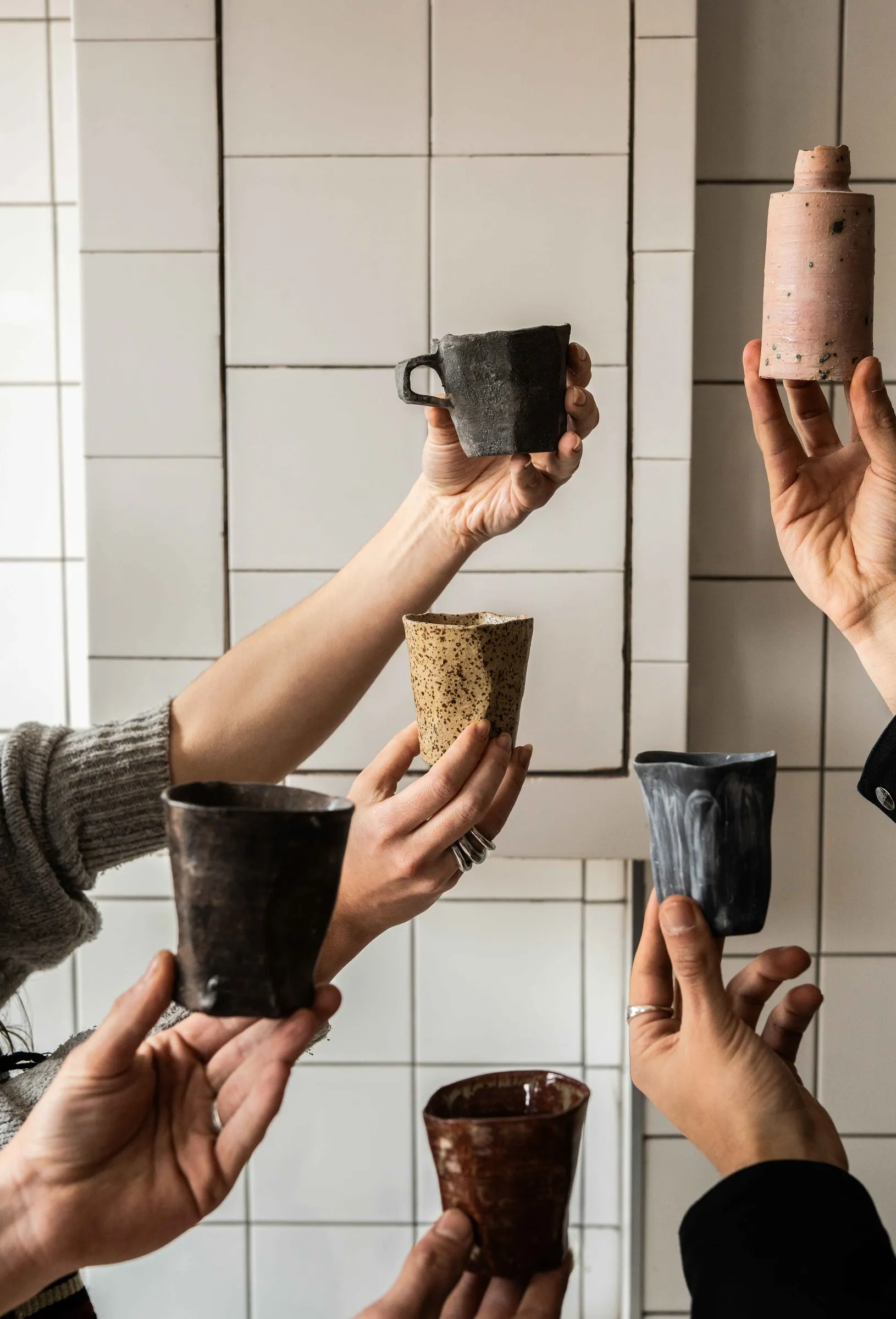 Several hands holding up handmade cups and a pink bottle against a white tiled wall