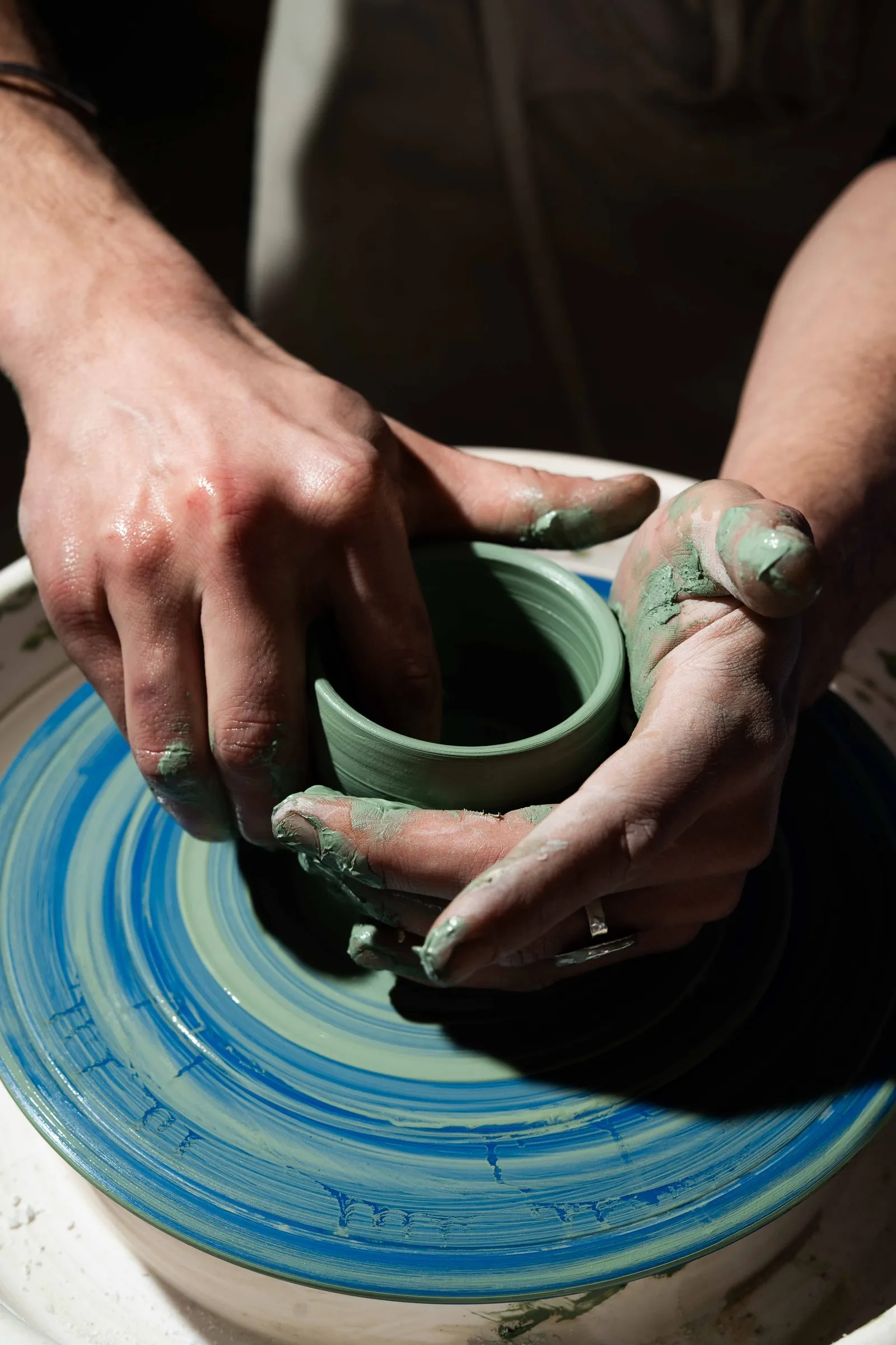 Clay-covered hands shaping a small green cup on a potter's wheel