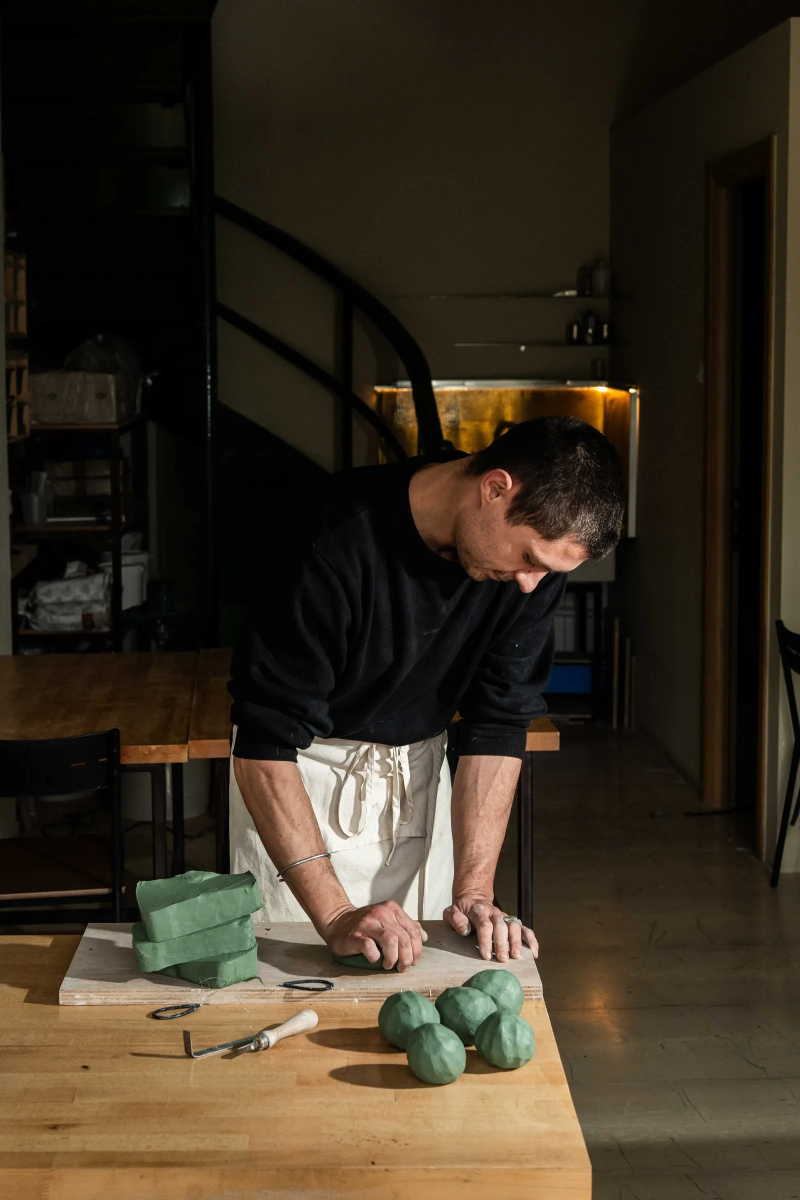 A ceramicist in an apron kneading green clay at a wooden table, clay balls beside him